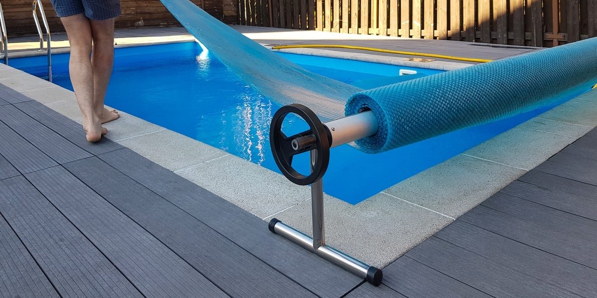 A barefoot man in blue shorts pulls a blue cover over a square pool enclosed by a brown wooden fence.