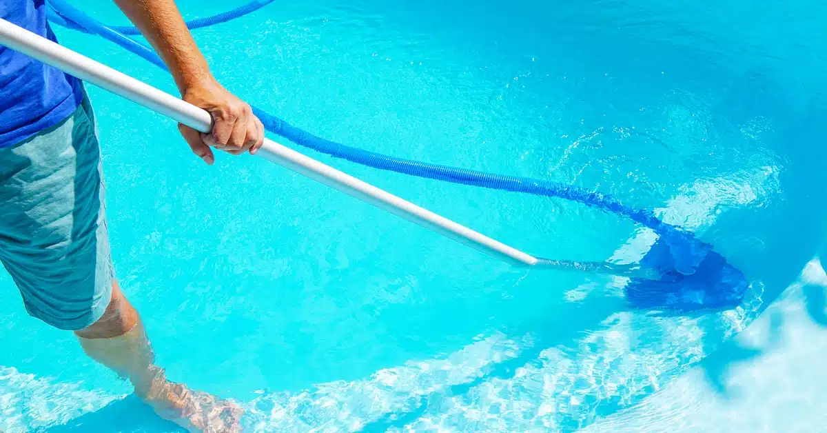 A person in a blue shirt and shorts uses a tool with a long hose to clean the bottom of a pool on a sunny day.