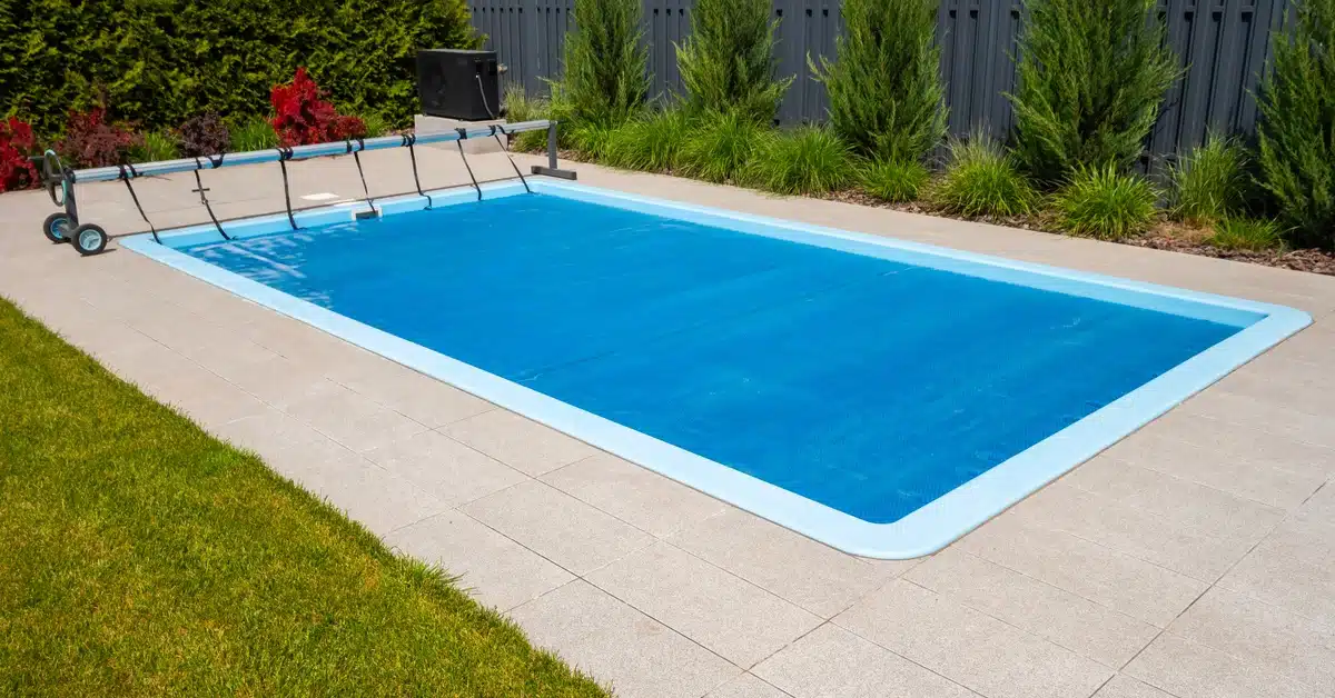 A rectangular outdoor pool with a blue cover surrounded by grass, landscaping, and a dark wooden fence.