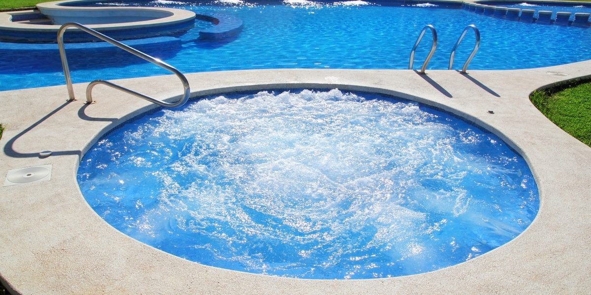 An outdoor circular spa with jets next to a large, curved pool, surrounded by short grass on a sunny day.