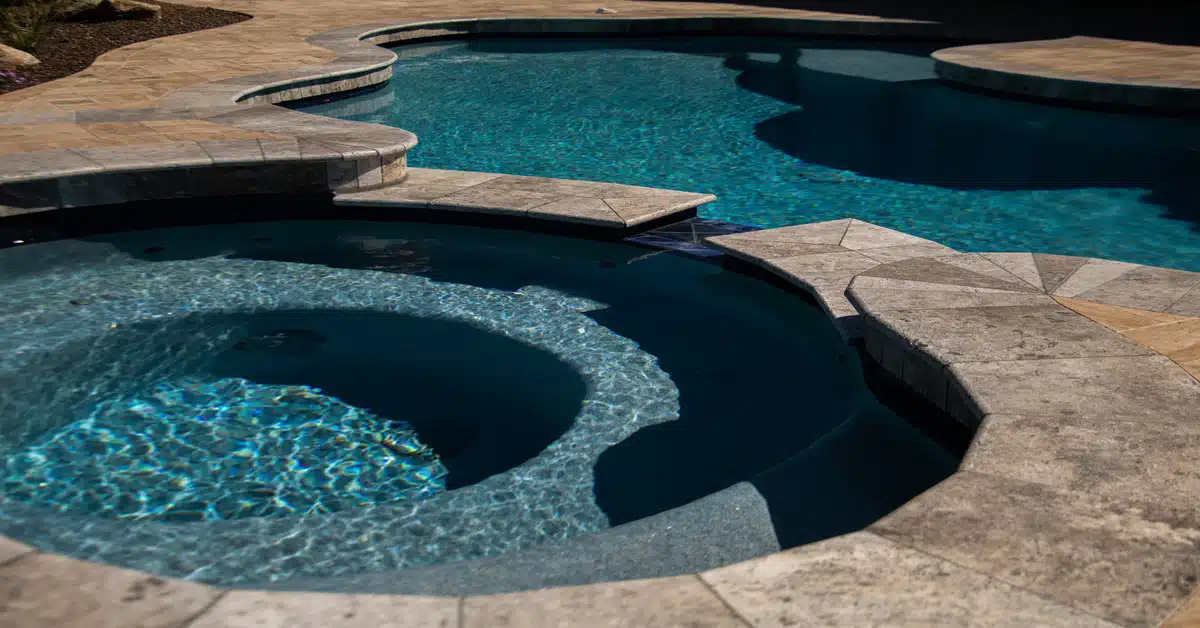 An outdoor circular spa connected to a curved pool surrounded by brown travertine tile on a sunny day.