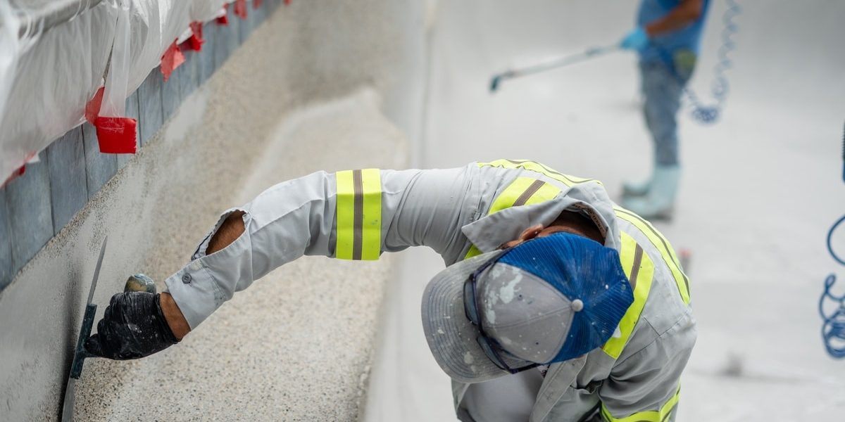 A worker in gray reflective gear, a hat, and black gloves uses a trowel to apply plaster to a textured pool surface.