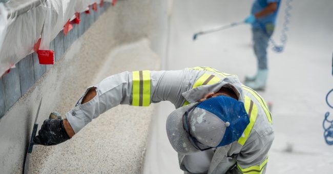 A worker in gray reflective gear, a hat, and black gloves uses a trowel to apply plaster to a textured pool surface.
