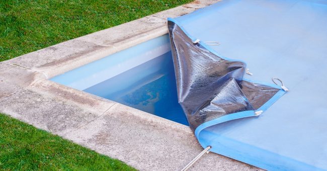 The corner of a swimming pool with a stone border, its blue cover partially peeled back to reveal the water.