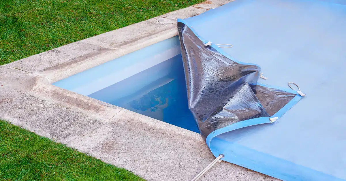The corner of a swimming pool with a stone border, its blue cover partially peeled back to reveal the water.