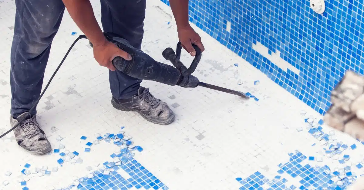 A worker uses a jackhammer to remove blue mosaic tiles from a pool floor, with broken tiles scattered across a white surface.