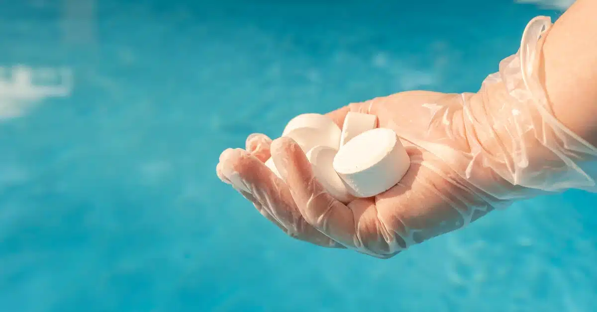 A gloved hand holding white chlorine tablets above clear blue pool water, ready for chemical treatment and sanitization.