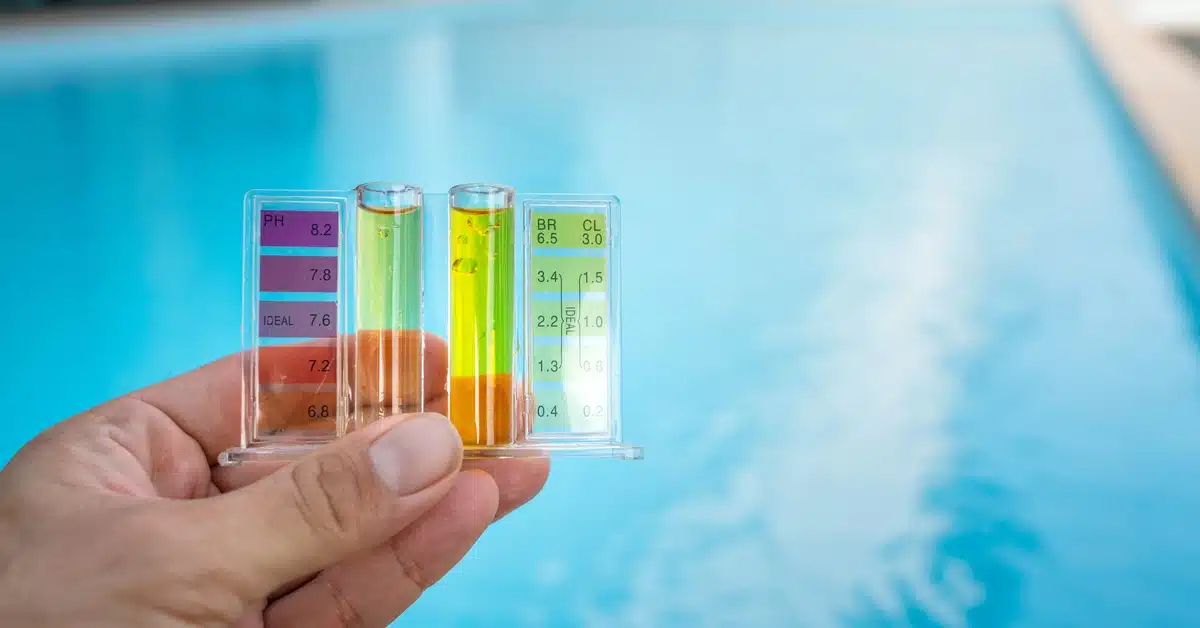 An individual holding a pool water test kit with colored chemical samples in front of a clear blue swimming pool.