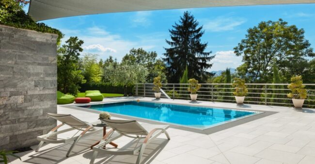A modern rectangular backyard pool with lounge chairs, tiled patio, railing, and lush trees under a covered terrace.