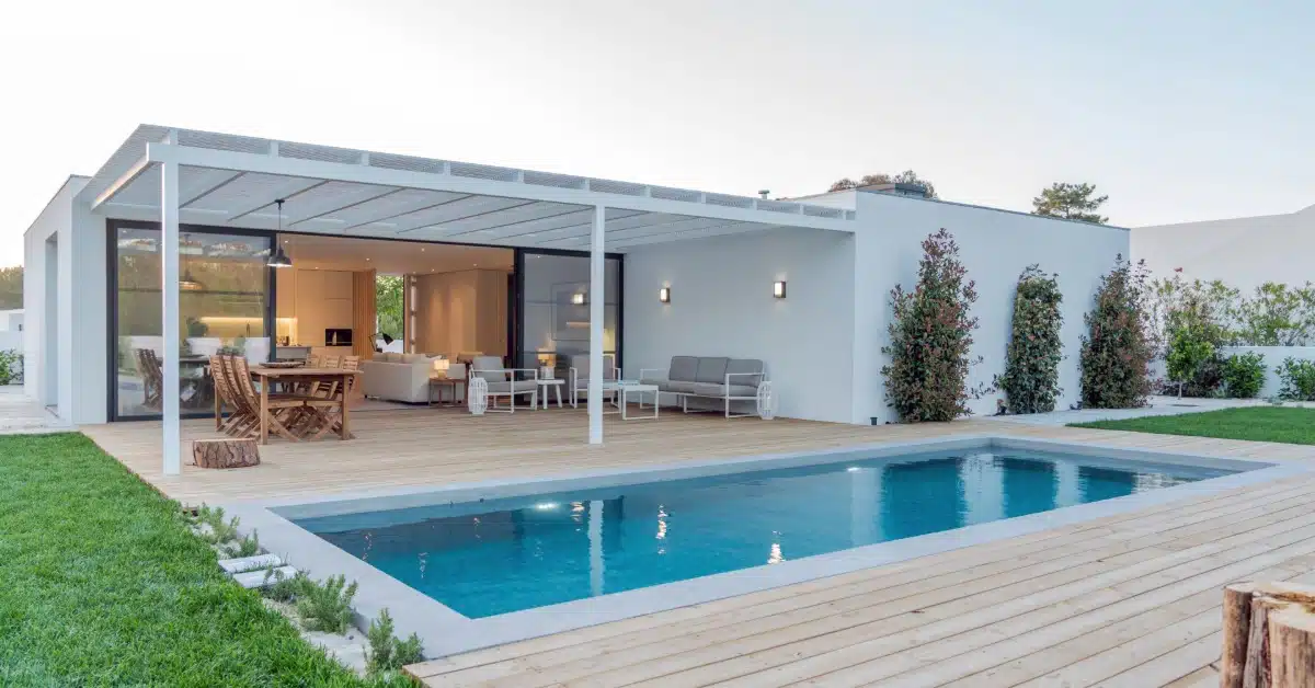A modern rectangular pool with a wood deck beside a white home and covered patio with outdoor seating.