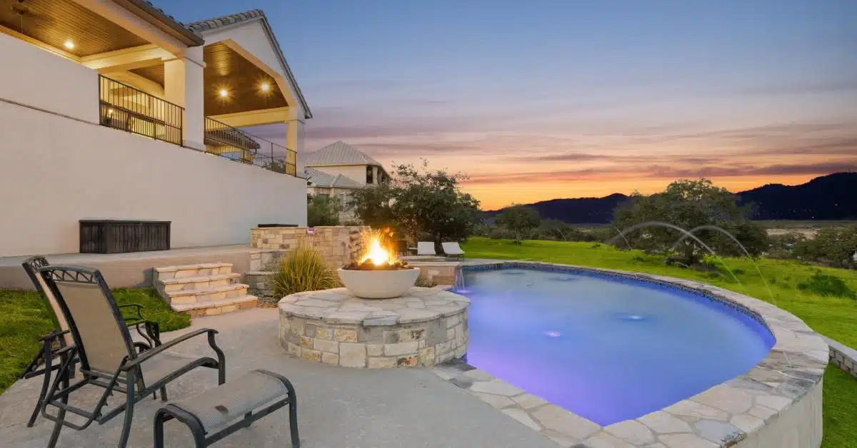A curved backyard pool at sunset with a raised stone fire bowl, patio chairs, and hillside view beside a large home.