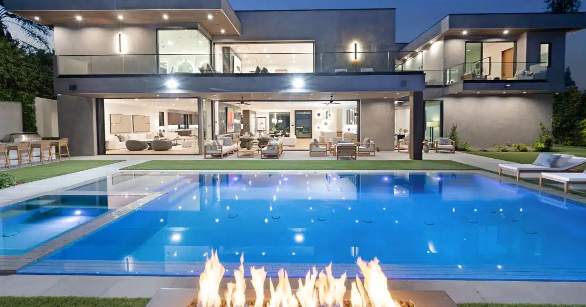 A modern luxury home with an illuminated backyard pool at night and a rectangular fire feature in the foreground.