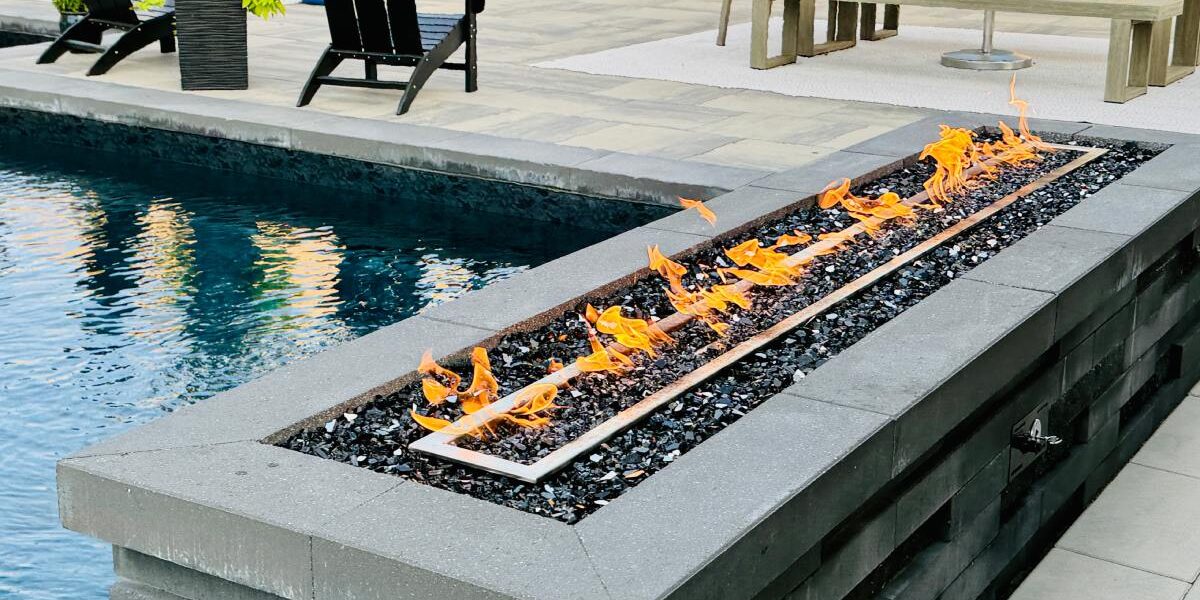 A long rectangular fire feature with orange flames set in stone beside a modern backyard pool, with patio chairs nearby.