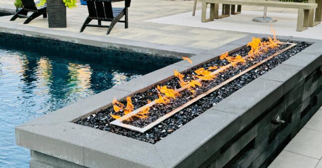 A long rectangular fire feature with orange flames set in stone beside a modern backyard pool, with patio chairs nearby.