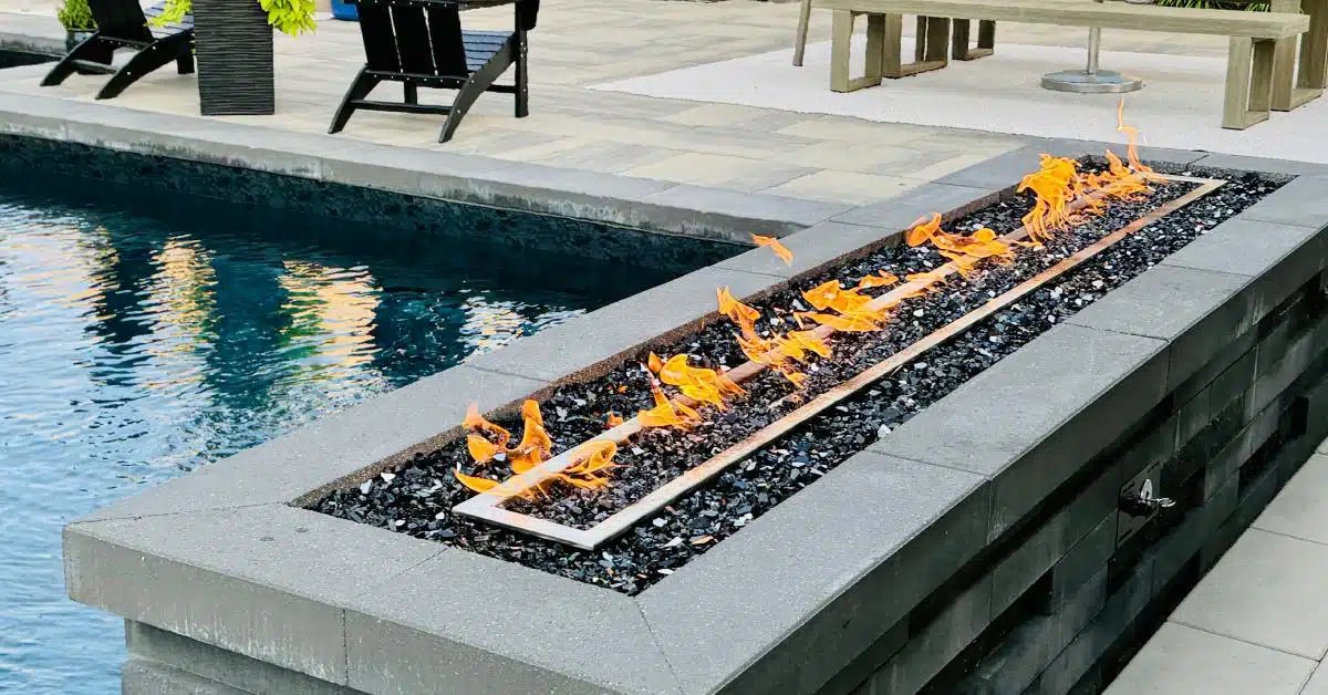 A long rectangular fire feature with orange flames set in stone beside a modern backyard pool, with patio chairs nearby.