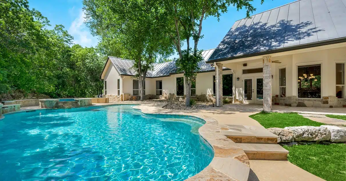 A freeform backyard pool with clear blue water beside a large home, featuring a stone deck and a shaded patio area.
