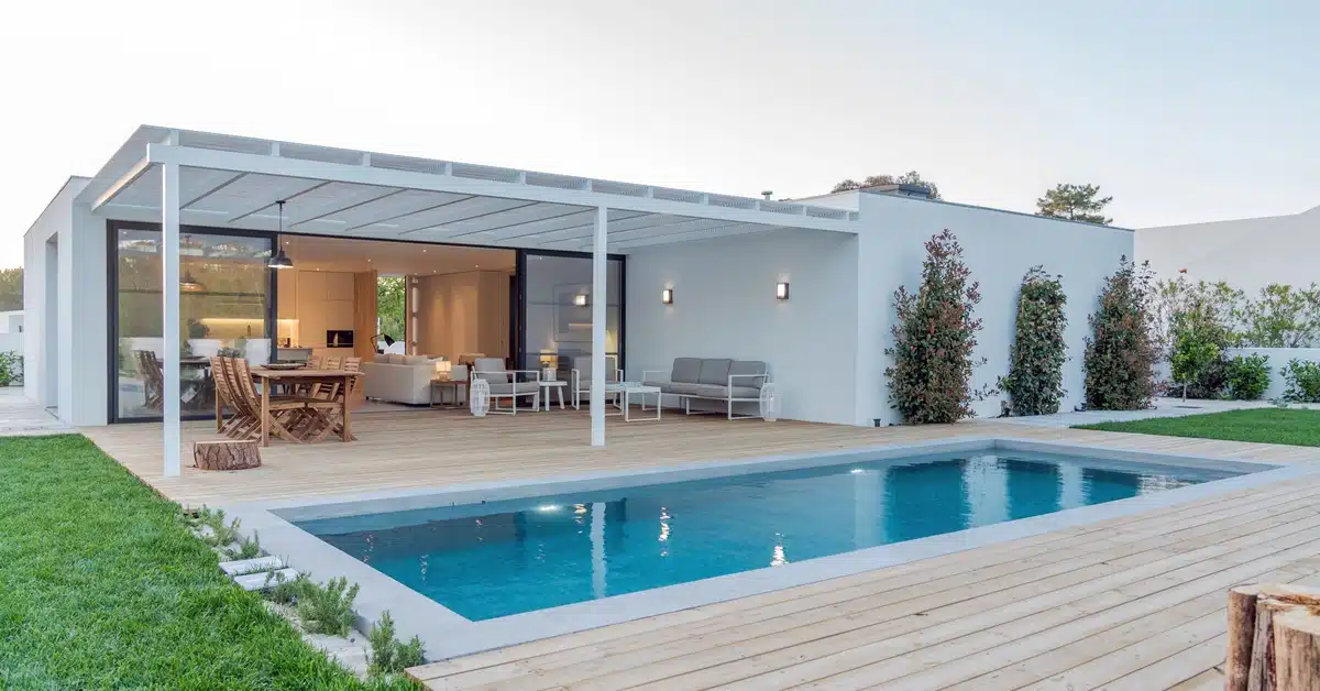 A modern white house with a rectangular pool beside a wooden deck and covered patio with outdoor seating.