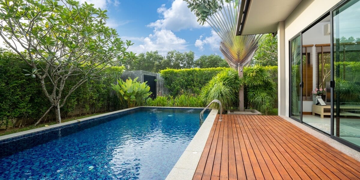A modern backyard pool with a wooden deck, surrounded by lush greenery and tall tropical plants under a blue sky. A modern backyard pool with a wooden deck, surrounded by lush greenery and tall tropical plants under a blue sky.