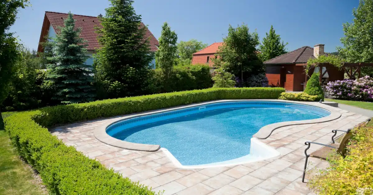 A backyard inground pool with a curved shape, surrounded by a paver deck, green hedges, trees, and nearby homes.