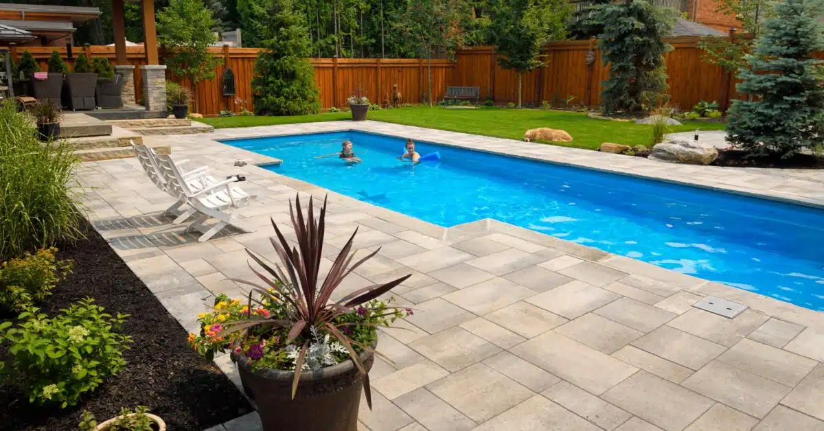 A rectangular backyard pool with two people swimming, surrounded by lounge chairs, a wooden fence, and a landscaped yard.