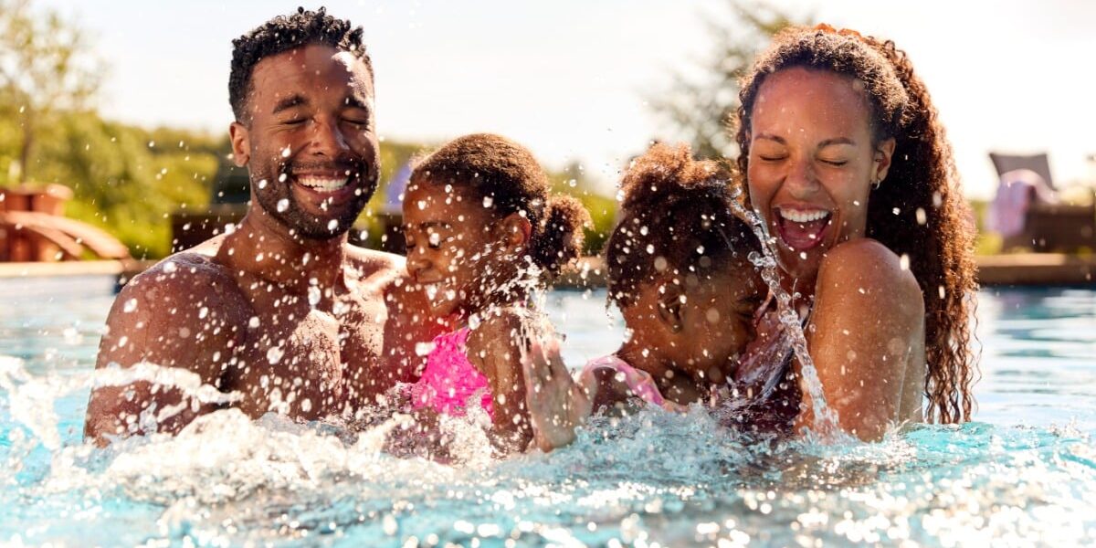 A smiling family with two young children splashing and laughing together in a bright outdoor swimming pool on a sunny day.