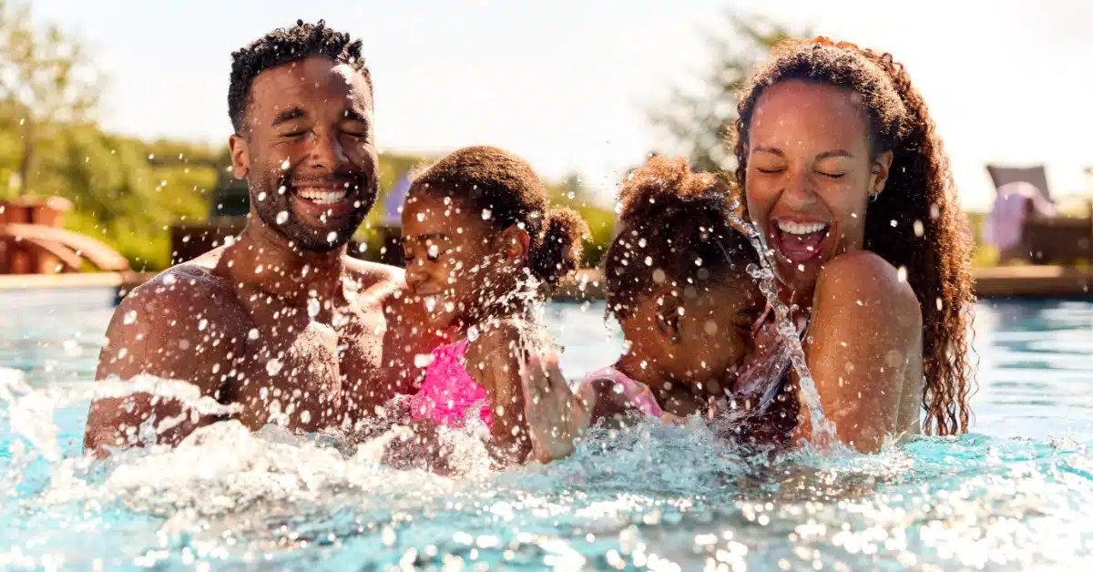 A smiling family with two young children splashing and laughing together in a bright outdoor swimming pool on a sunny day.