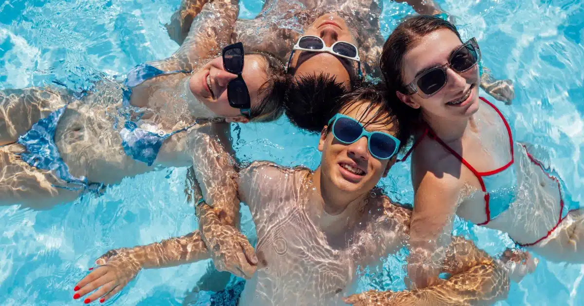 A group of friends wearing sunglasses smiling and floating together in a clear blue swimming pool on a bright sunny day.