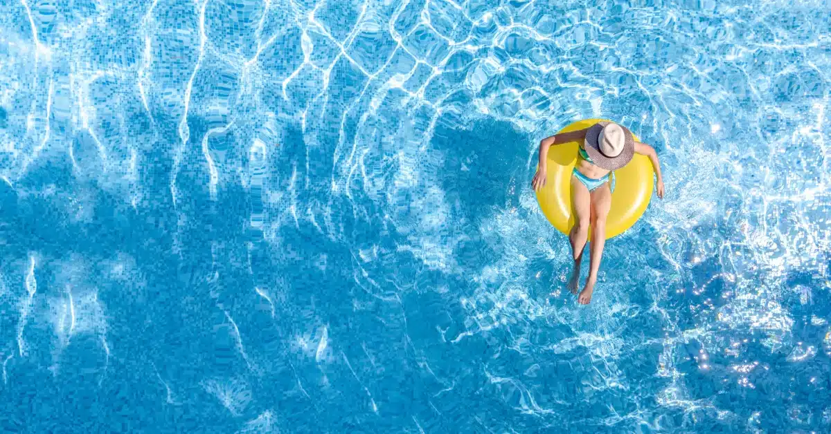 An overhead view of a woman relaxing on a yellow pool float, wearing a sunhat in clear blue water on a sunny day.
