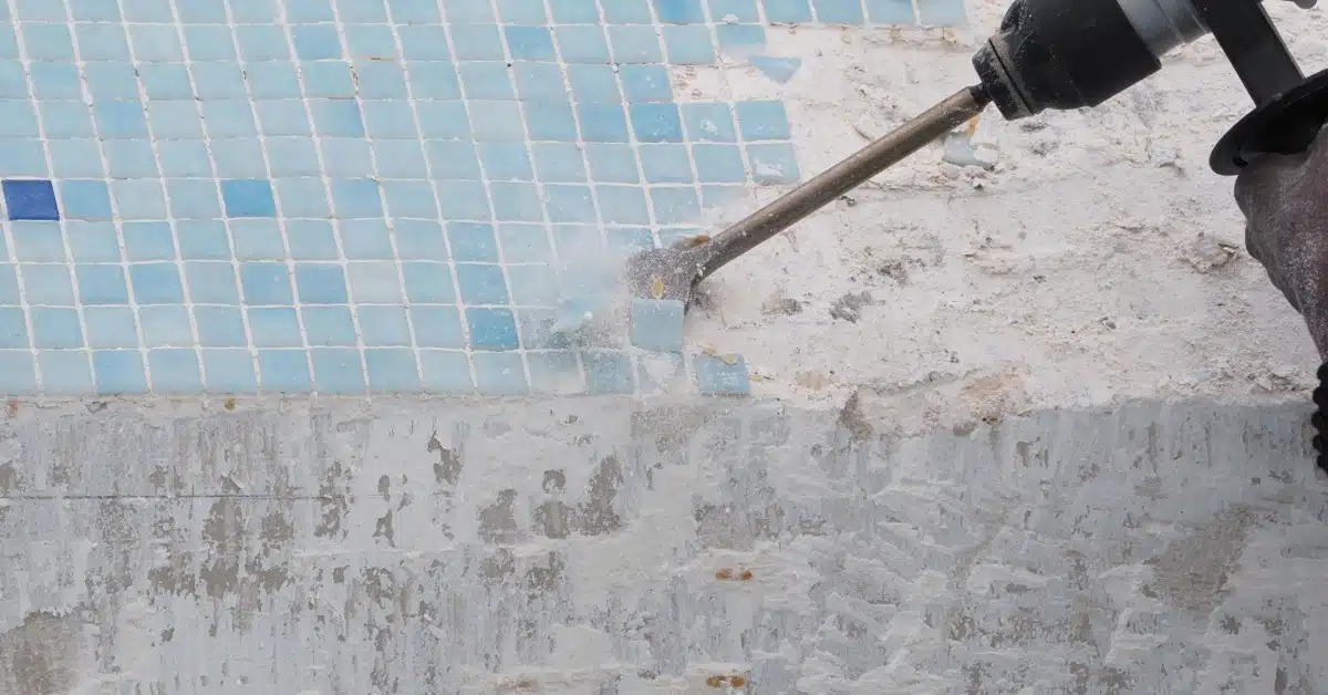 A power chisel removing blue pool tiles from a worn concrete surface during a swimming pool renovation process.