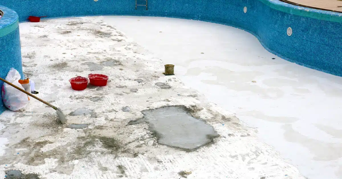 A drained swimming pool with a worn, damaged floor, visible patch repairs, and tools and buckets placed around the surface.