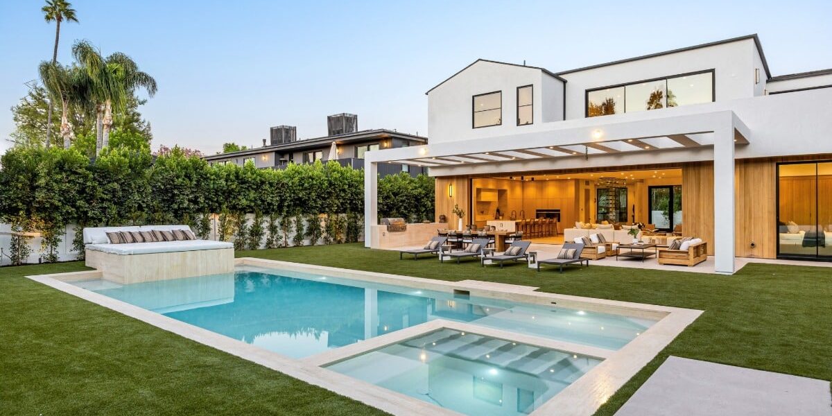 A modern white home with a geometric pool, raised tanning ledge, and a covered outdoor lounge surrounded by palm trees.