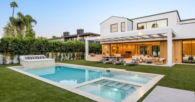 A modern white home with a geometric pool, raised tanning ledge, and a covered outdoor lounge surrounded by palm trees.