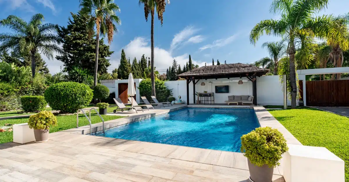 A backyard pool with tropical palm trees, lounge chairs, and a shaded outdoor cabana on a sunny day.